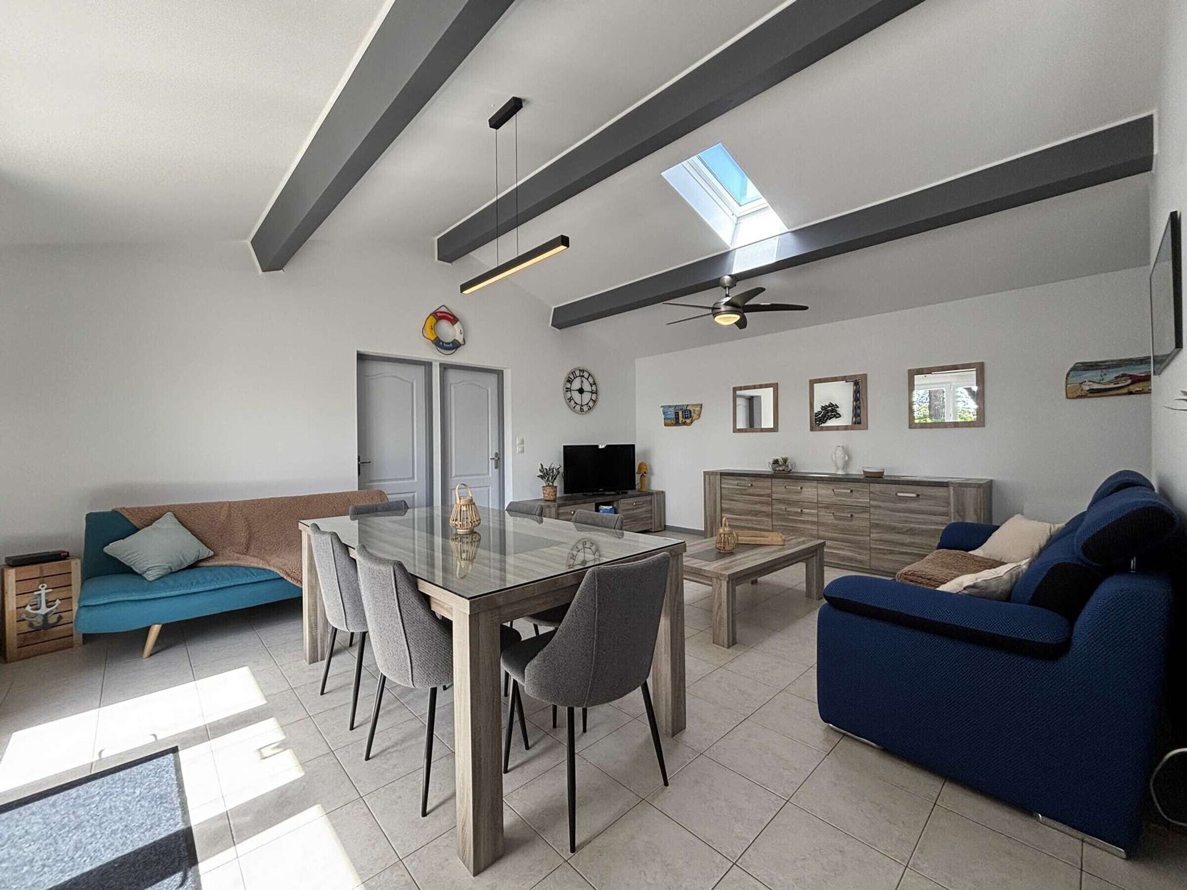 A modern living room with a glass dining table, grey sofas, a wooden cabinet, and a ceiling fan.