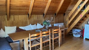 Ett matbord och stolar i trä, en soffa och en trappa i ett rum med träväggar och ett sluttande tak.