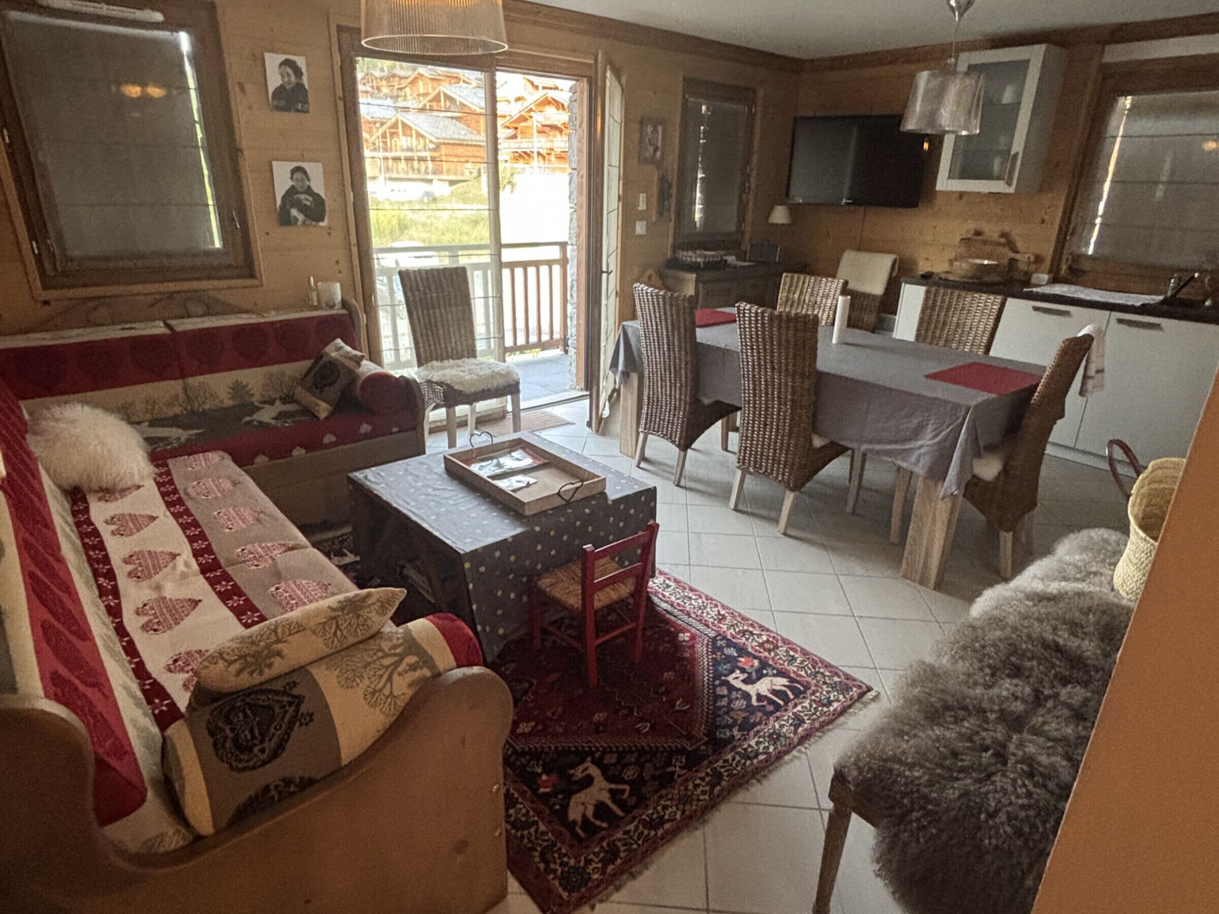 Een gezellige woonkamer met een bank, een salontafel en een vloerkleed. Er is een eethoek met stoelen en een tafel. De keuken is zichtbaar, met kastjes en een televisie.