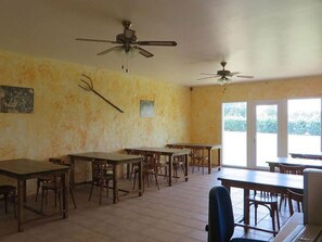 Une pièce spacieuse, dotée de plusieurs tables et chaises, de ventilateurs de plafond et de grandes fenêtres laissant entrer la lumière naturelle.