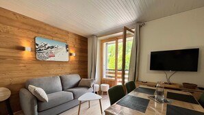 Moderni olohuone, jossa on harmaa sohva, pieni valkoinen pöytä, puinen ruokapöytä tuoleineen, seinälle asennettu televisio ja suuri ikkuna, jossa on verhot.