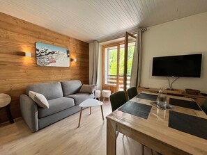 Ein modernes Wohnzimmer mit grauem Sofa, einem kleinen weißen Tisch, einem Holzesstzimmer mit Stühlen, einem an der Wand montierten Fernseher und einem großen Fenster mit Vorhängen.