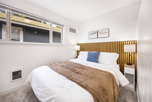 Ein Schlafzimmer mit Bett, Kopfstütze, Nachttisch mit Lampe und einem Fenster mit Jalousien.