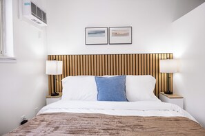A hotel room with a bed, two bedside lamps, a wooden headboard, and two framed pictures on the wall. - Modern Workstay in Seattle | Desk + Full Kitchen (Seattle)