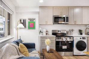 A modern kitchen with a microwave, oven, and washing machine. There is a sofa with cushions, a lamp, and a vase with dried flowers on a wooden table. - Modern Workstay in Seattle | Desk + Full Kitchen (Seattle)