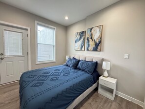 Ein Schlafzimmer mit einem Bett, einem Nachttisch, einer Lampe, einer Tür und zwei an der Wand befestigten Bildern.