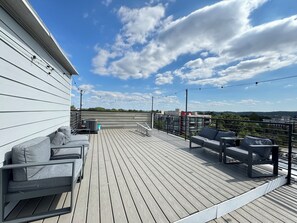 Eine Holzterrasse mit Sitzgelegenheiten im Freien, einem Geländer und freiem Blick auf den Himmel und entfernte Gebäude.