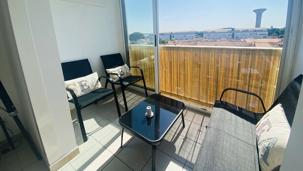 A balcony with a glass door, two chairs, a small table, and a view of buildings and a tower. - Studio cabin, parking, near beach - 4 guests (Le Grau-du-Roi)