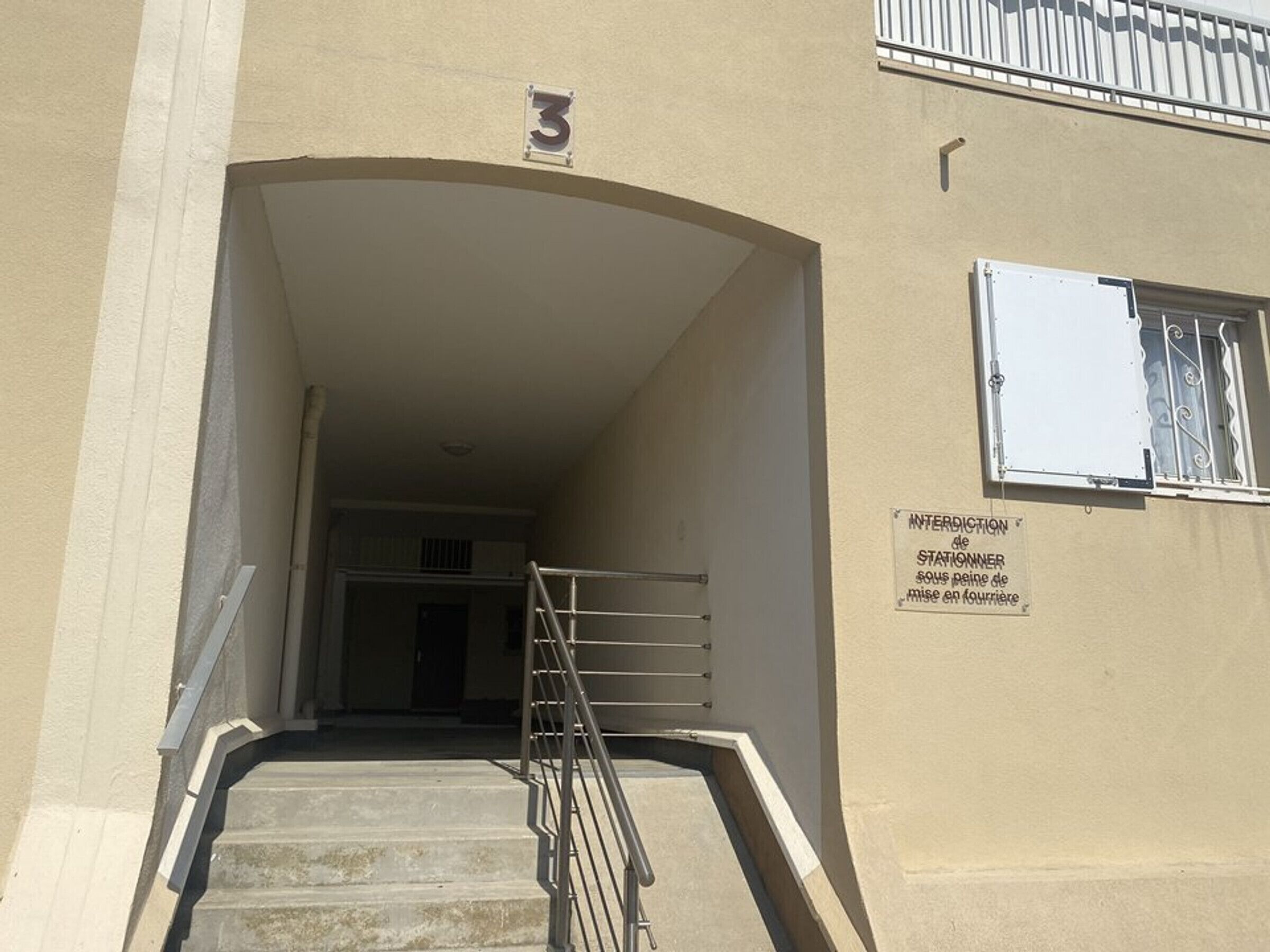 Une entrée d’immeuble avec un escalier menant à une porte, une plaque portant le numéro « 3 » au-dessus de la porte, et une fenêtre avec une inscription en français.