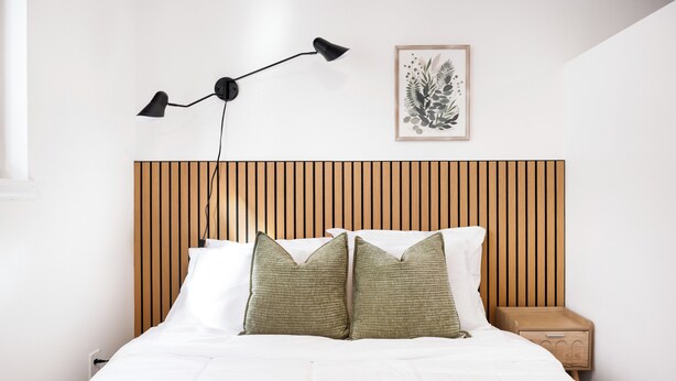 Ein ordentlich bezogenes Bett mit weißen Leinen und zwei Kissen, ein Bettgestell aus Holzlamellen, eine Nachttischlampe, ein gerahmtes Pflanzenbild und ein Nachttisch mit einer Schublade.