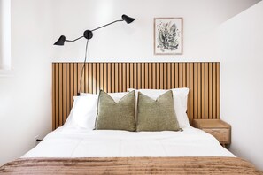 Un lit bien fait, avec du linge de lit blanc et deux oreillers, une tête de lit en lattes de bois, une lampe de chevet, une estampe botanique encadrée et une table de chevet en bois avec un tiroir.