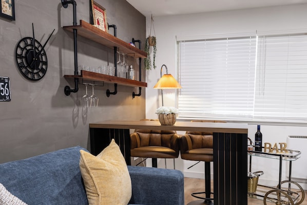 A modern living room with a blue sofa, two leather bar stools, a wooden coffee table, a wall-mounted clock, shelves with glassware, and a window with blinds. - Modern Industrial Seattle Suite | Workspace + Kitchen (Seattle)