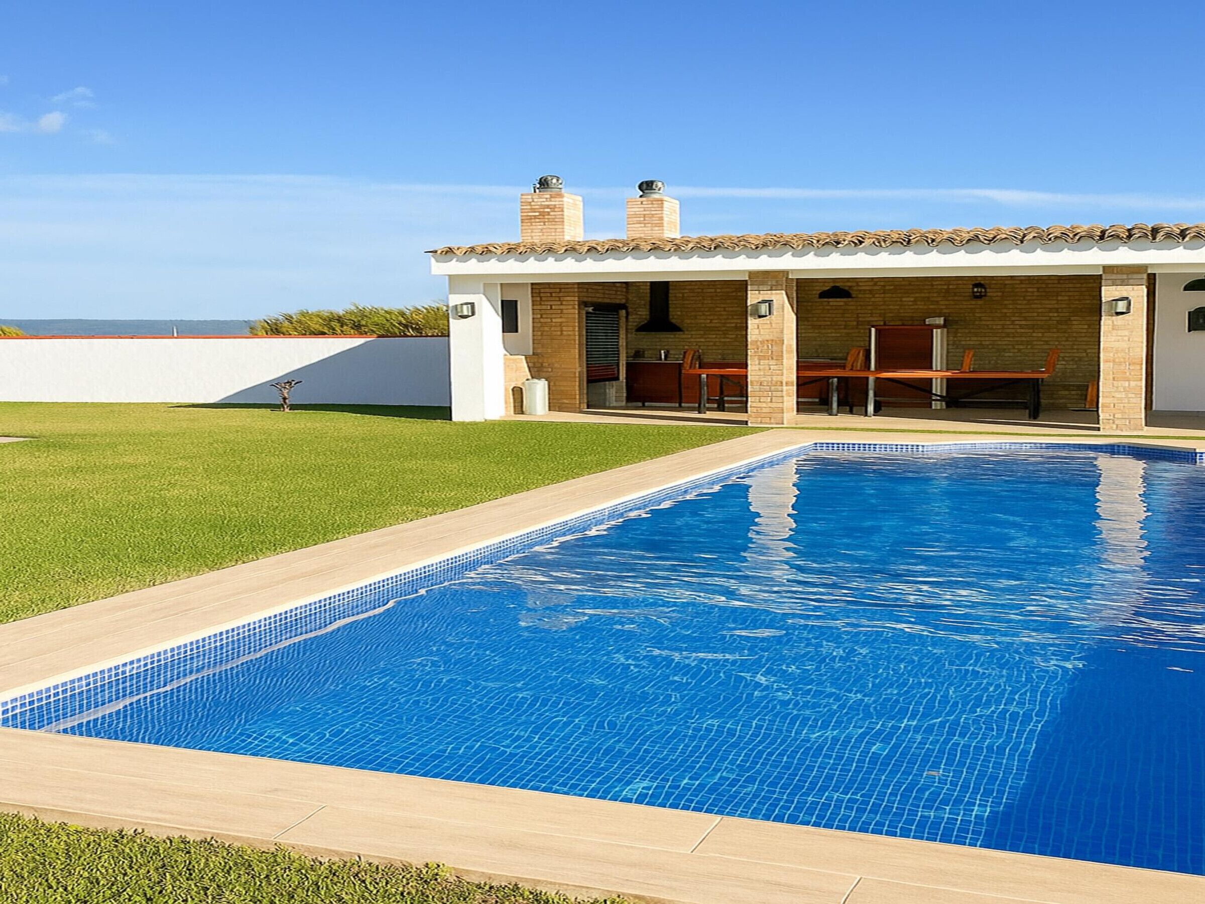 Piscina com borda em azulejo, área de terraço com mobiliário externo e um edifício com telhado em azulejo e paredes de tijolo.
