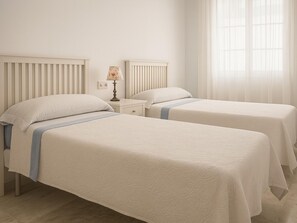Een tweeepersoonskamer met twee bedden, een nachtkastje, een lamp en een raam met dunne gordijnen.