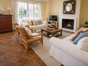 Ein Wohnzimmer mit Kamin, weißen Sofas, Korbstühlen, einem Couchtisch und einem Fernseher.