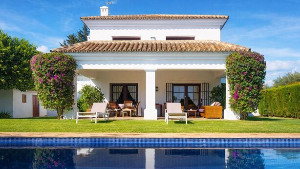 Ein zweistöckiges Haus mit Swimmingpool, einer überdachten Terrasse mit Gartenmöbeln und einem Garten mit blühenden Pflanzen.