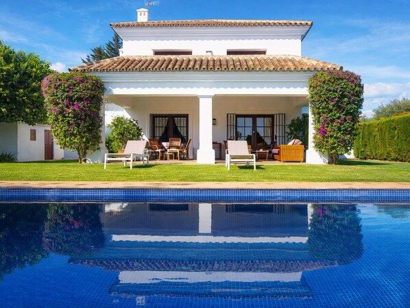 Ein zweistöckiges Haus mit Swimmingpool, einer überdachten Terrasse mit Gartenmöbeln und einem Garten mit blühenden Pflanzen.