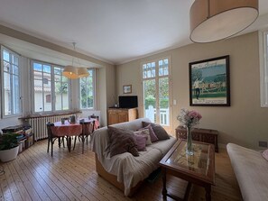 A cozy room with a wooden floor, a sofa, a dining table with chairs, a TV, and a painting on the wall. - Large typical Bauloise villa - 12 guests (La Baule)