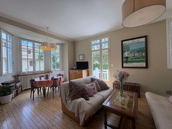 A cozy room with a wooden floor, a sofa, a dining table with chairs, a TV, and a painting on the wall.
