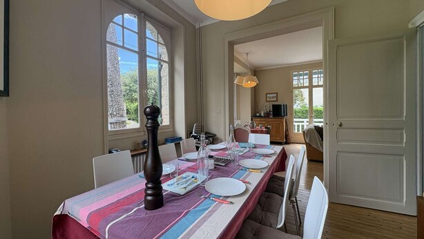 Une salle à manger avec une longue table dressée pour quatre personnes, de grandes fenêtres et une vue sur l’extérieur.