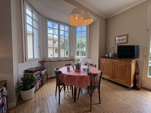 A dining area with a table, chairs, a radiator, a bookshelf, a potted plant, and a television on a wooden cabinet.