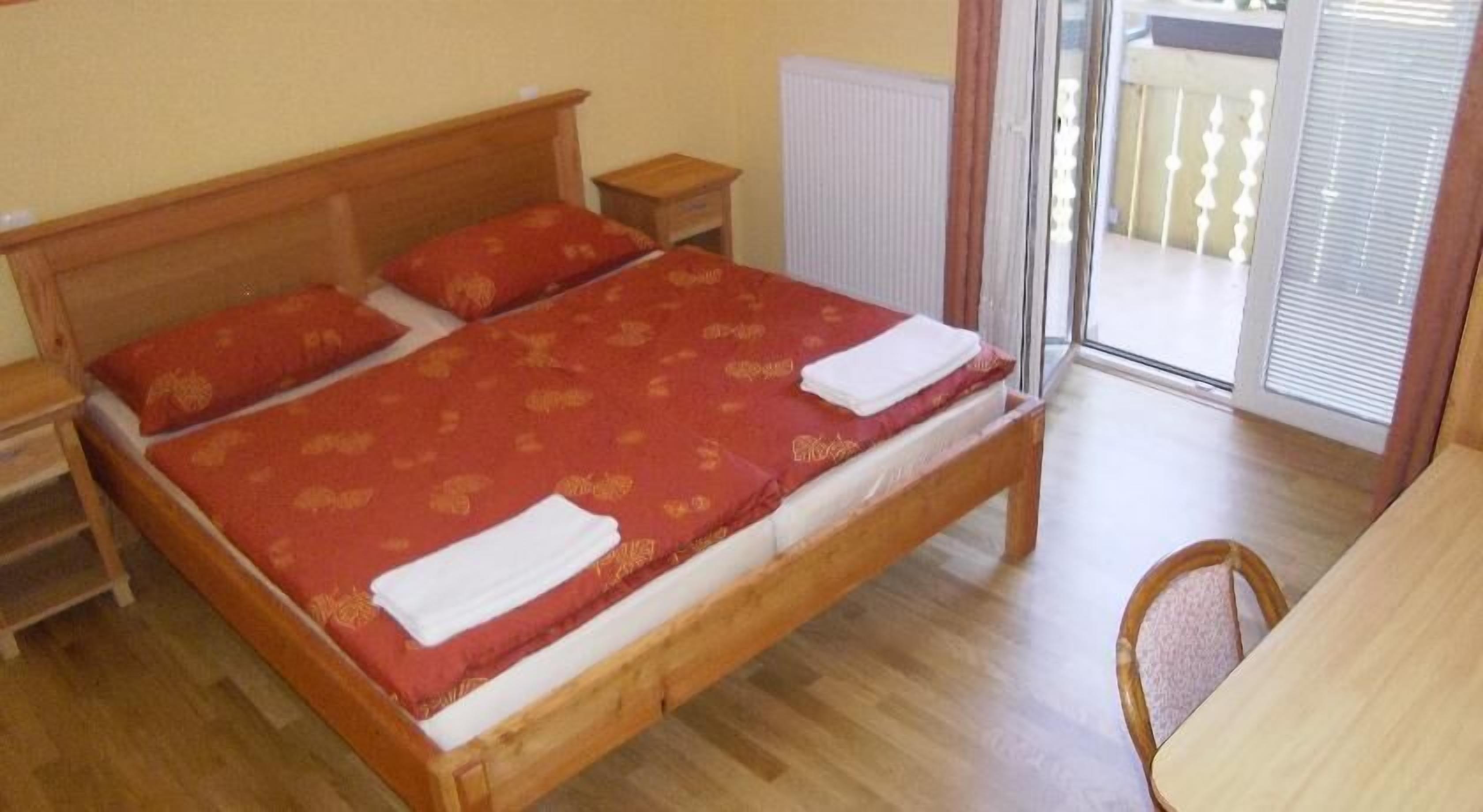 Quarto com cama de madeira, mesa de cabeceira, cadeira e vista para o exterior através de uma porta de vidro.