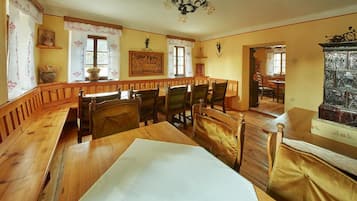 Uma área de refeições com bancos e cadeiras de madeira, um candelabro e vista para a sala de jantar através de uma janela.
