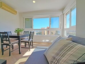 Ein Zimmer mit Esstisch, Stühlen, einer Couch und großen Fenstern mit Blick auf Gebäude.