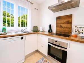 Et moderne køkken med hvide skabe, en opvaskemaskine, ovn, emhænde i rustfrit stål og et vindue med udsigt til grønne områder.