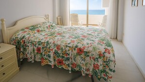 Un dormitorio con cama, mesita de noche, silla, balcón con vista al mar y aire acondicionado.