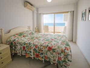 A bedroom with a bed, a nightstand, a chair, a balcony with a view of the sea, and an air conditioning unit. - Apartment with Parking in Estepona, by the Sea (Estepona)