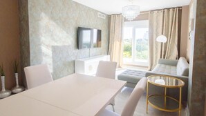 Een moderne woonkamer met een witte eettafel, een bank, een aan de muur bevestigde televisie en een groot raam met gordijnen.
