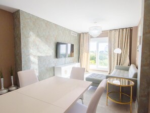 A modern living room with a white dining table, a sofa, a TV mounted on the wall, and a large window with curtains. - Luxury Apartment in Estepona with Terrace and Parking (Estepona)
