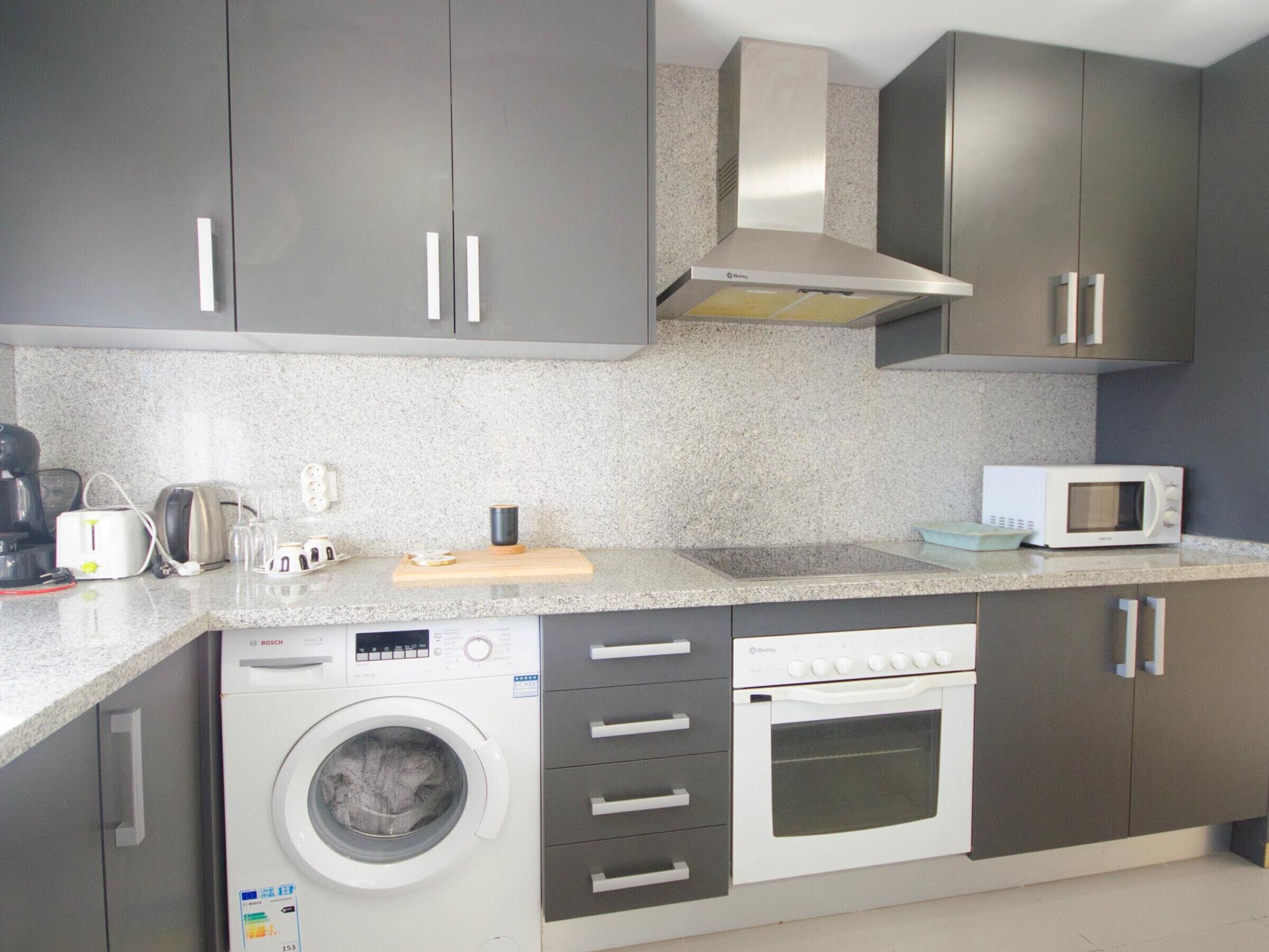 Cocina compacta con armarios grises, lavadora Bosch, microondas y tostadora.