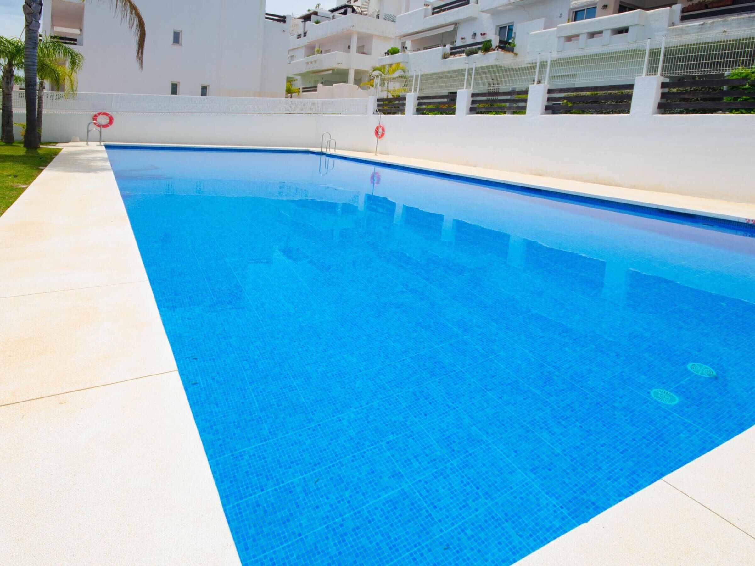 En rektangulär swimmingpool med kristallklart blått vatten, omgiven av ett vitt poolområde och en vit vägg med balkonger.