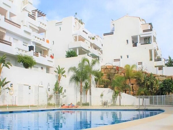 A pool area with white buildings, palm trees, and a rocky outcrop. - Luxury Apartment in Estepona with Terrace and Parking (Estepona)