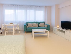 Un saló modern amb un sofà color turquesa, una taula de cafè blanca, un televisor sobre un moble blanc i una zona de menjador amb taula i cadires blanques.