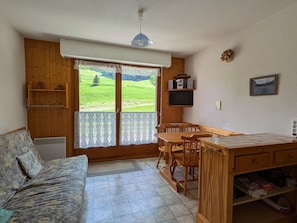 Ein gemütliches Zimmer mit einer Holzsofa, einem Esstisch mit Stühlen, einem Fernseher und Blick auf eine grüne Landschaft durch die Tür.