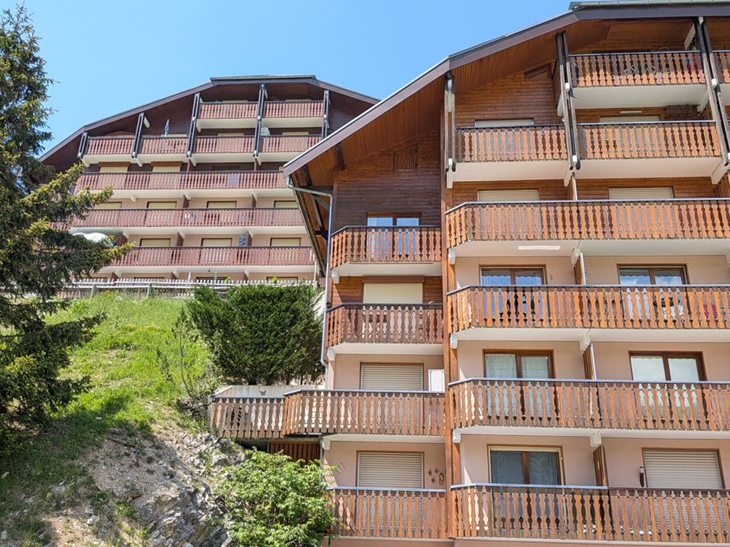 A multi-story building with balconies, a sloped roof, and a hillside background.