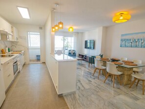 Eine moderne Küche mit weißen Schränken, einem Essbereich mit Glastisch und einem an der Wand befestigten Fernseher.