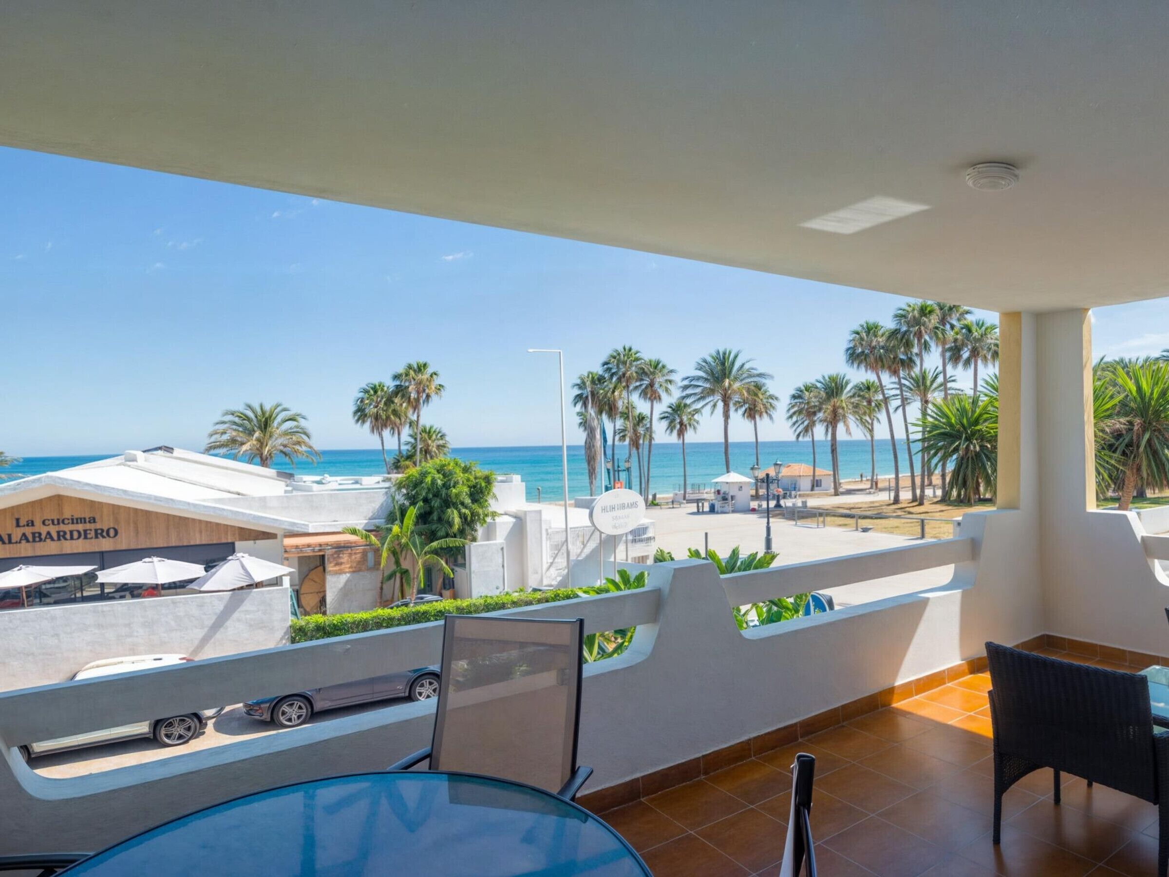 Uma varanda com mesa de vidro, cadeiras e vista para a praia e palmeiras.