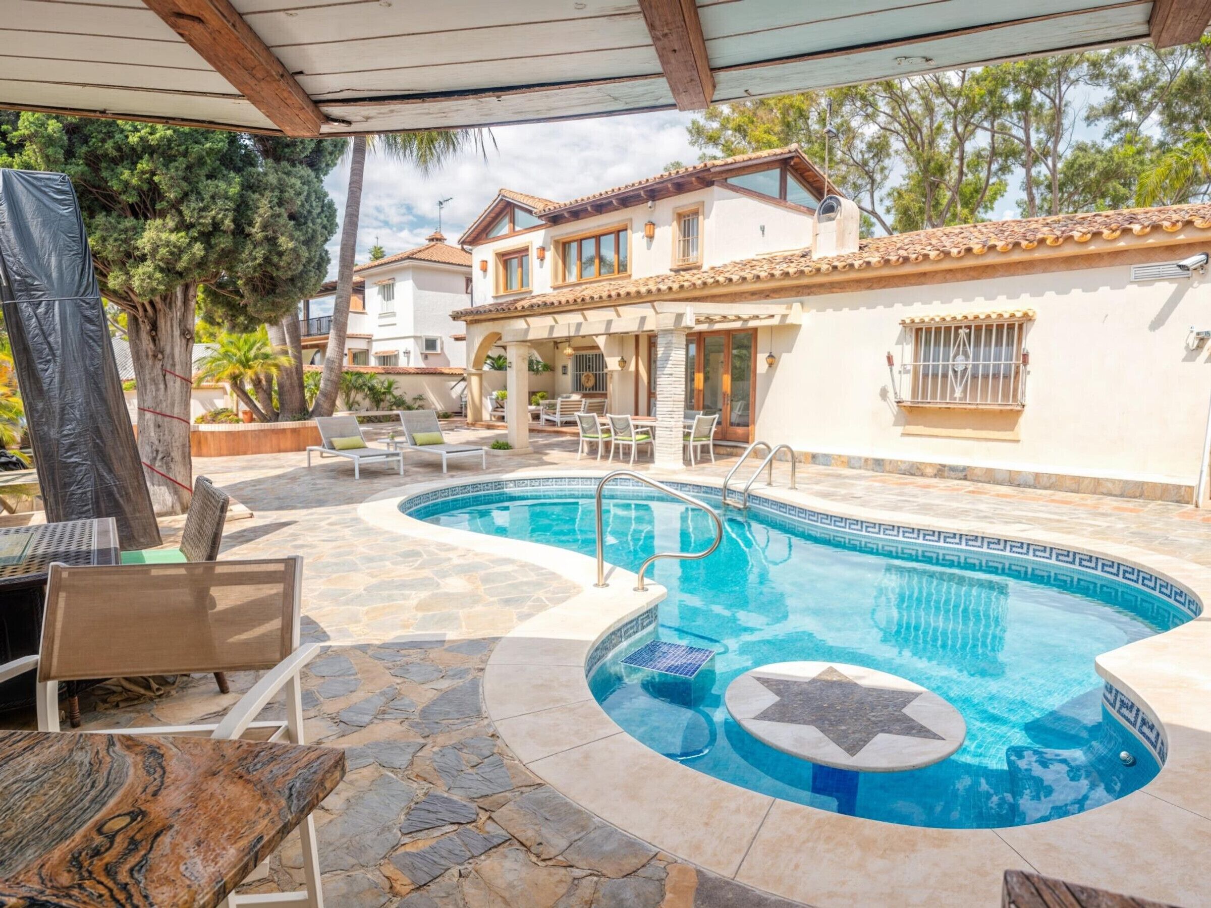 A swimming pool with a star-shaped design, surrounded by stone tiles and a covered patio area with lounge chairs.