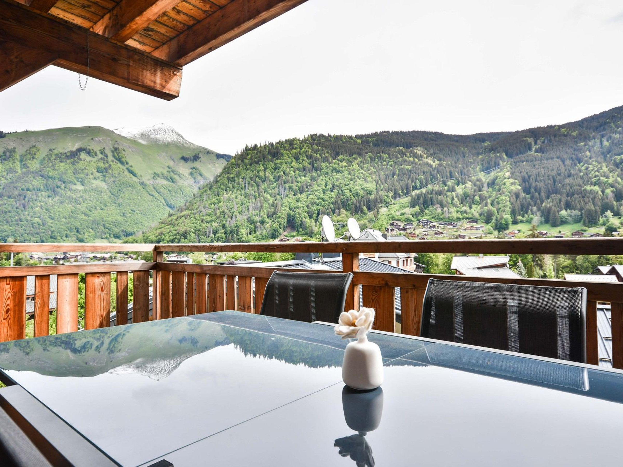 A wooden balcony with a glass table and chairs, overlooking a mountainous landscape.