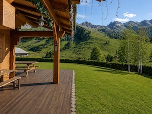 Una terraza de madera con bancos, una pérgola con luces colgantes y vistas a una ladera verde con montañas al fondo.