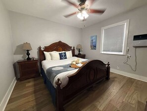 Ein Schlafzimmer mit einem hölzernen Bett, zwei Nachttischen mit Lampen, einem Deckenventilator, einem Fenster mit Jalousien und einem Bild an der Wand.