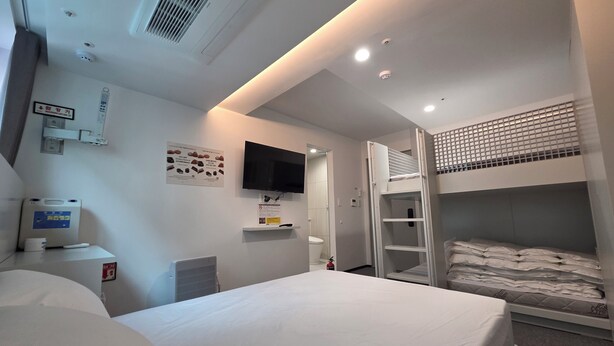 Una habitación de hotel con cama, televisor, litera, estante pequeño y aire acondicionado instalado en la pared.