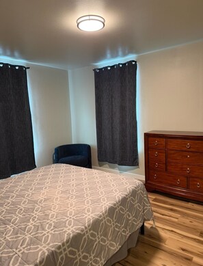 A bedroom with a bed, a dresser, a chair, and two windows with curtains.