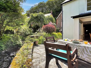 A stone pathway leading to a wooden bench with a table set for breakfast, surrounded by lush greenery and a stone building. - Glen Cottage, Porlock (Porlock)