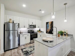 Cozinha moderna com armários brancos, eletrodomésticos de aço inoxidável, bancadas de granito e iluminação pendente.