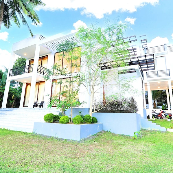A modern two-story building with balconies, a garden area with small trees, and a clear sky.